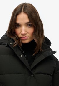 Sort pufferjakke med høj krave, trykknapper og lynlåse detaljer. Blød tekstur og quiltet mønster er fremtrædende.