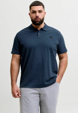 Mężczyzna z brodą, ubrany w granatową koszulkę polo i jasnoszare spodnie, stoi na jednolitym tle.