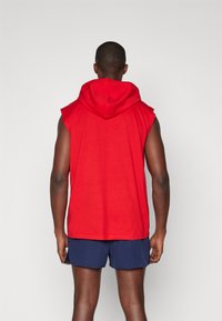 Red sleeveless hoodie with a large hood and soft texture, paired with navy athletic shorts. Back view, showcasing hoodie design and fit.