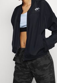 Svart bombarjacka med ribbade manschetter och krage, med en subtil Nike Air-logotyp. Bärs över en ljusblå cropped top med svarta detaljer.