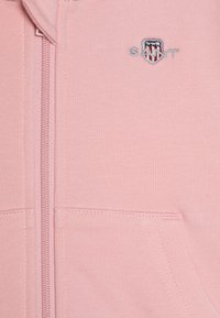 Felpa con zip di colore rosa chiaro, realizzata in tessuto morbido, con tasche cucite e un logo ricamato sul petto. Texture liscia con un design casual.