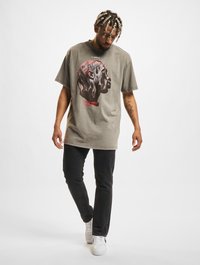 Graues, oversized T-Shirt mit einem markanten Grafikdruck eines Gesichts, kurzen Ärmeln und einem runden Ausschnitt, kombiniert mit schwarzen Jeans und weißen Schuhen.