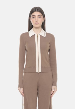 Donna con capelli castani lisci che indossa un cardigan marrone con zip e pantaloni abbinati, con colletto bianco e strisce laterali.