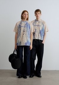 Μπλουζάκια σε συνδυασμό μπεζ και μπλε με γραφικό σχέδιο, συνδυασμένα με σκούρα τζιν με φαρδιά μπατζάκια. Ένα μοντέλο κρατάει μια μαύρη τσάντα.