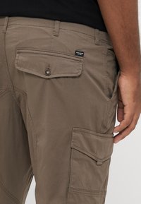 Καφέ παντελόνι cargo με πίσω τσέπη με φλάπα και κουμπί. Κατασκευασμένο από ανθεκτικό ύφασμα, με εμφανείς ραφές και ετικέτα.