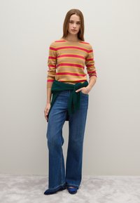 Gestreifte Strickjacke in Beige, Pink und Gelb, kombiniert mit blauen weit geschnittenen Jeans. Ein grüner Pullover, der um die Taille gebunden ist, fügt einen Kontrast hinzu. Blaue Schuhe.