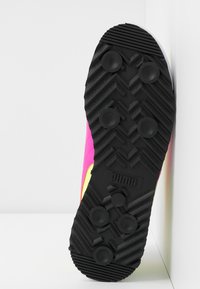 Semelle noire à crampons d'une chaussure de sport Puma avec tige rose et jaune, positionnée verticalement contre un mur blanc dans un coin.