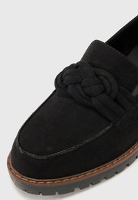 Mocasin de ante negro con acento anudado, suela plana y detalles de costura. Presenta una plantilla acolchada y suela de goma para mayor agarre.