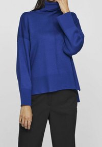 Maglione dolcevita blu cobalto con vestibilità ampia, polsini a costine e orlo posteriore leggermente più lungo, abbinato a pantaloni neri. Tessuto liscio.