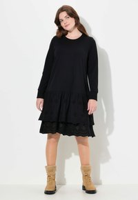 Robe noire à manches longues avec un design à œillets superposés en bas, associée à des bottes à doublure en fourrure tan et un col rond.