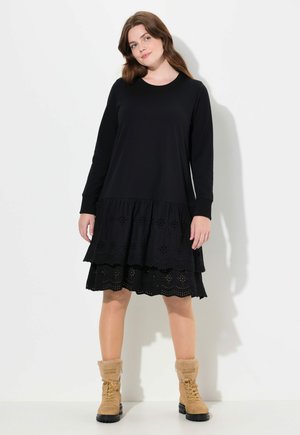 Schwarzes Langarmkleid mit mehrlagigem Lochmuster am Saum, kombiniert mit tanfarbenen, mit Fell gefütterten Stiefeln und einem runden Ausschnitt.