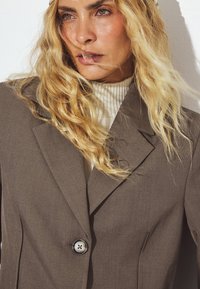 Blazer marrone su misura con rever a scollo e chiusura a un bottone. Indossato su un dolcevita a coste chiaro. Textura morbida, dettagli minimi.