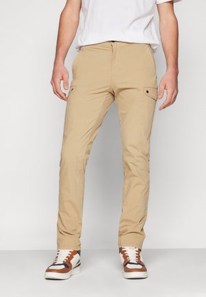 Pantaloni cargo beige con una vestibilità dritta, dotati di tasche laterali e dettagli con bottoni. Realizzati in tessuto leggero, abbinati a sneaker bianche e marroni.