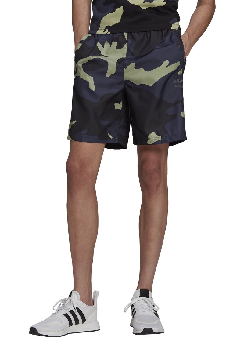 Pantaloni corti con stampa camouflage nera e verde, dotati di una cintura elastica, tasche laterali e un tessuto liscio e leggero. Indossati con scarpe da ginnastica bianche.