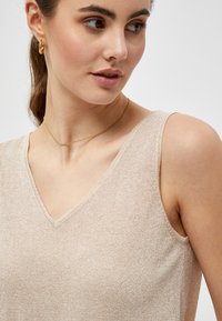 Beige sleeveless top featuring a V-neck design, made from textured fabric with a subtle shimmer. Gold necklace and hoop earring accents.
