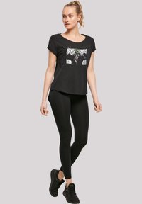Svart t-skjorte med grafikktrykk på forsiden, kombinert med svarte tights. Modellen har på seg svarte joggesko. Glatt stoff, avslappet passform.