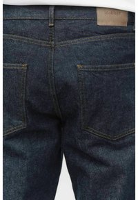 Mörkblå denimjeans med en slät textur, med två bakfickor och kontrasterande gul sömnad. En tan-färgad märkesetikett finns ovanför midjan.