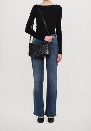 Black crossbody bag with embossed pattern and silver hardware. Model wearing fitted black top, flared blue jeans, and black flats.