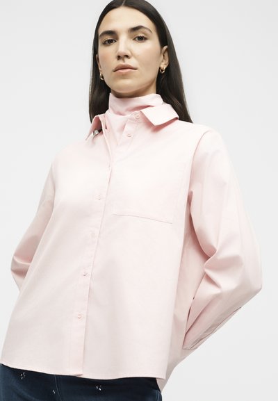 Γυναίκα με μακριά σκούρα μαλλιά φοράει ανοιχτό ροζ πουκάμισο με κουμπιά και αντίστοιχο ζιβάγκο, στέκεται με τα χέρια πίσω από την πλάτη της σε απλό φόντο.