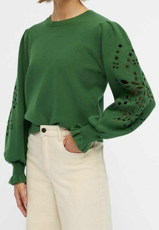 Pull vert à manches bouffantes avec des motifs floraux découpés brodés, associé à un pantalon beige clair. Texture douce, col rond.