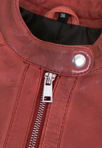 Rote Lederjacke mit Nahaufnahme eines silbernen Reißverschlusses und Druckknopfs, mit strukturiertem Finish und sichtbaren Nähten.