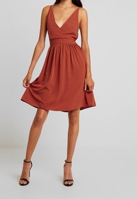 Šaty A-line v rustové barvě s hlubokým výstřihem do V a tenkými ramínky. Mají přiléhavý pas a splývavou sukni, vyrobené z lehkého, hladkého materiálu.