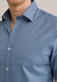 Camisa de botón azul claro, con una textura suave y mate, cuello puntiagudo y botones blancos contrastantes. Tejido de algodón, corte ajustado.