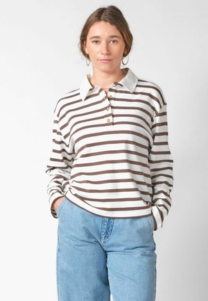 Kvinde med brunt hår bærer store hoop-øreringe, en hvid og brunstribet langærmet polo og lyseblå jeans med hænderne i lommerne.