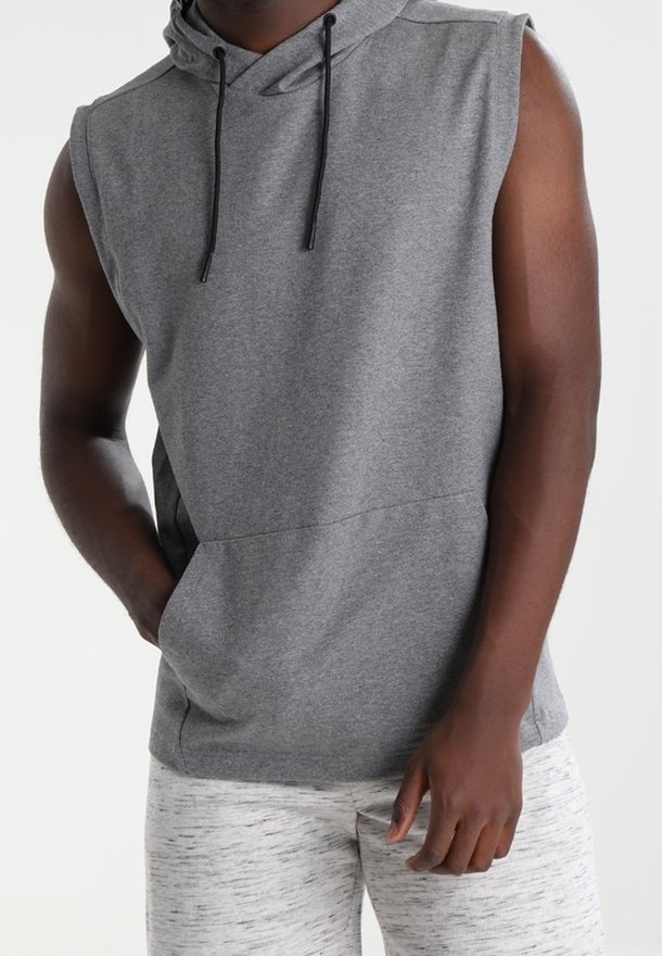 Persona vestida con un hoodie sin mangas gris y pantalones de chándal gris claro, de pie con una mano en el bolsillo contra un fondo liso.