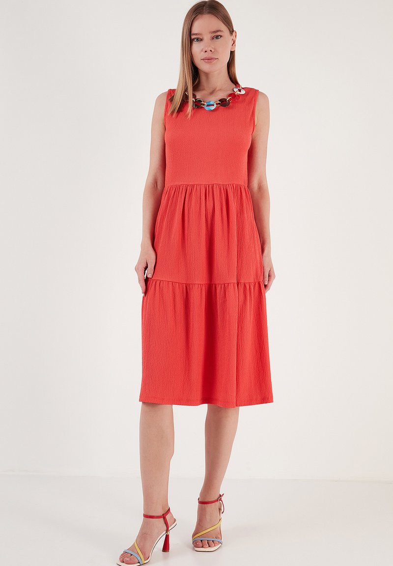 Ärmelloses, knielanges Kleid in leuchtendem Orange mit strukturiertem Stoff und gestuftem Design. Akzentuiert mit einer bunten Halskette.