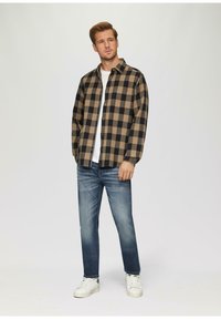 Geruite overhemd in zwart en beige, van een katoenmix, met knoopsluiting; gecombineerd met lichtblauwe jeans en witte sneakers. Model staat tegen een neutrale achtergrond.