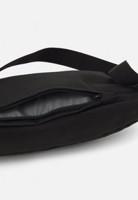 Czarna torba materiałowa o teksturowanej powierzchni, z zamykaną na zamek komorą i gładkim wykończeniem wnętrza. W zestawie regulowany pasek.