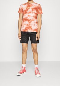 Camiseta naranja de tie-dye con "WORLD TOUR" en letras doradas, combinada con pantalones cortos de mezclilla negros y zapatillas de caña alta rosas.