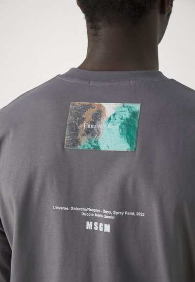 Grijze t-shirt met een textuurpatch met abstracte groene en bruine patronen op de achterkant; gedrukt tekstdetails onder de patch in het wit.