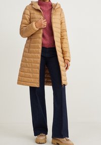 Abrigo acolchado color marrón claro con capucha y costuras horizontales. Combinado con un suéter rosa y vaqueros de pierna ancha azul oscuro.