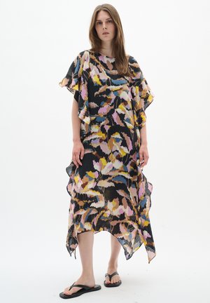 Mujer con un vestido suelto hasta la rodilla con un estampado floral abstracto multicolor en negro, rosa, amarillo y azul, combinado con sandalias negras.
