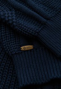 Granatowy sweter z dzianiny o teksturowym wzorze. Posiada brązową metkę z wygrawerowanym napisem "OMBRE HERITAGE" przymocowaną do materiału.