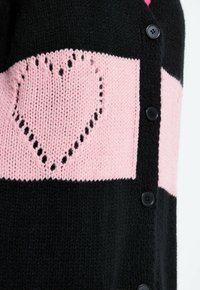 Czarny sweter na drutach z szerokimi różowymi poziomymi paskami i wzorem serca w różowym pasku, zapięty na czarne guziki na przodzie.