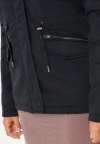Giacca nera realizzata in un tessuto robusto, con due tasche con zip e dettagli con cordino. L'orlo è dritto e presenta un aspetto texturizzato.