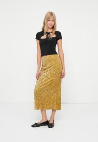 Una donna bionda sta in piedi indossando una maglietta nera a maniche corte con nodo frontale e una lunga gonna di paillettes dorate con ballerine nere su uno sfondo bianco.