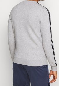 Sweatshirt gris avec des poignets et un ourlet côtelés, présentant des rayures latérales noires avec des détails blancs. Texture douce, coupe décontractée, encolure ronde.