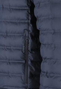Nærbillede af en mørkeblå quilted jakke, der viser vandrette syninger og en lodret lynlås-lomme.