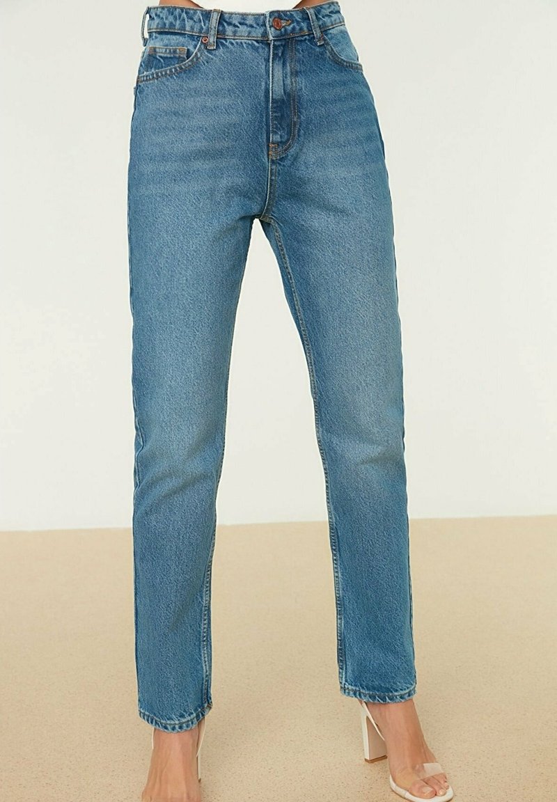 Jean bleu taille haute à jambe droite porté avec des sandales à talon bloc et bride transparente sur un sol beige.