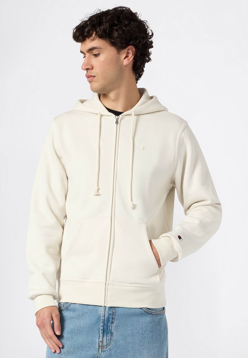 Haut à capuche crème zippé avec une poche avant, des cordons de serrage et un accent de logo sur la poitrine ; associé à un jean en denim bleu clair. Texture de tissu douce.