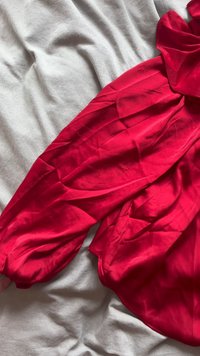 Rode blouse met lange mouwen en een textuurafwerking, met pofmouwen en een losse pasvorm. De stof ligt op een lichtgrijze ondergrond.