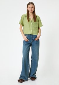Camisa corta de manga larga texturada y verde con cuello y botones, combinada con jeans anchos azules y zapatos de plataforma marrones.