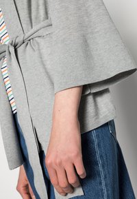 Persona con un cárdigan gris con cinturón sobre una camisa a rayas y vaqueros azules, con un enfoque en los detalles de la mano izquierda y la manga.