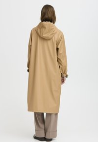 Imperméable beige avec capuche, manches longues et coupe décontractée. Présente une doublure intérieure à motifs et un design long jusqu'aux genoux.