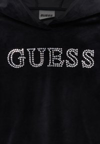 Crna baršunasta kapuljača s "GUESS" logotipom izrađenim od srebrnih rhinestona, ističe glatku teksturu i sjajne detalje.