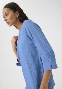 Haut bleu clair en tissu texturé avec des manches trois-quarts, présentant des fentes latérales et une coupe ample. Apparence douce et froissée.